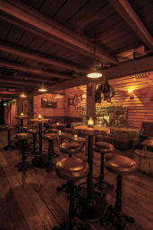 Cozy rustic cabin bar interior with wood-paneled walls and exposed beams, stone fireplace, round high-top tables, leather stools, and warm amber lighting.