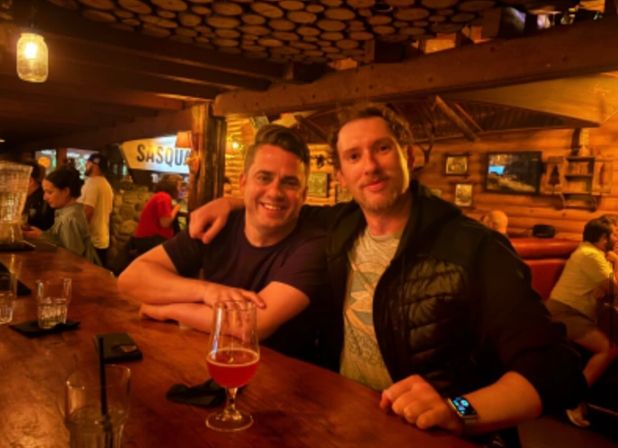 Two people smiling at a wooden bar in a cozy log-cabin style pub, warm amber lighting, a beer glass on the counter and rustic wood decor in the background.