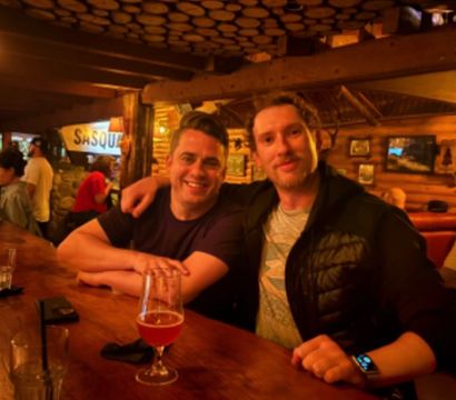 Two people smiling at a wooden bar in a cozy log-cabin style pub, warm amber lighting, a beer glass on the counter and rustic wood decor in the background.
