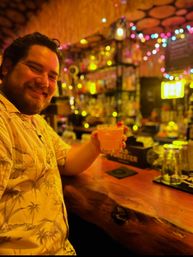 Smiling man in a Hawaiian shirt holding a tropical cocktail with a cherry at a dim, tiki-style bar—warm yellow lighting, wooden counter, and colorful string lights bokeh in the background.