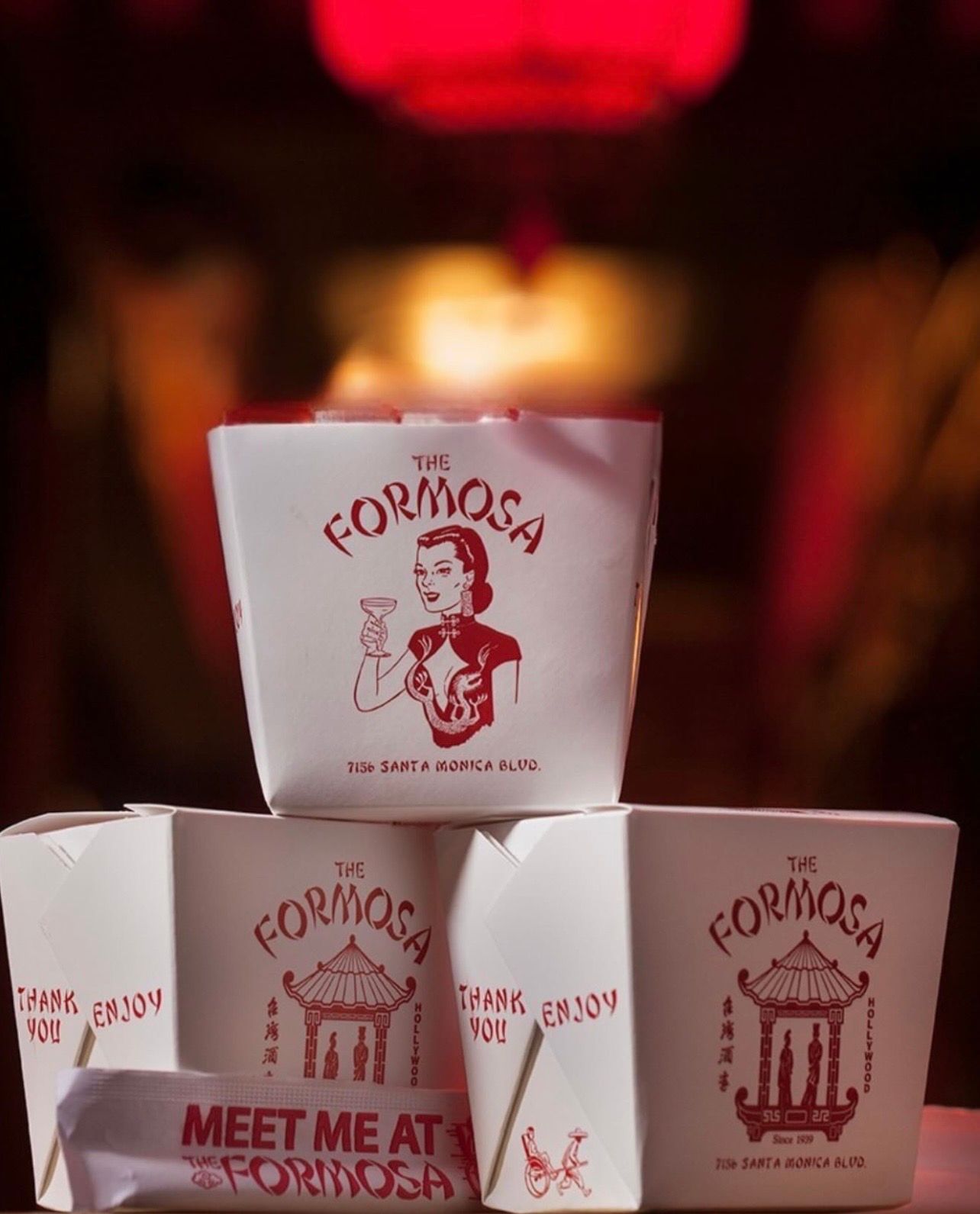 Stack of red-and-white Chinese takeout boxes with vintage-style illustrations and a 'Meet me at' napkin, bathed in moody red restaurant lighting