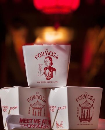 Stack of red-and-white Chinese takeout boxes with vintage-style illustrations and a 'Meet me at' napkin, bathed in moody red restaurant lighting
