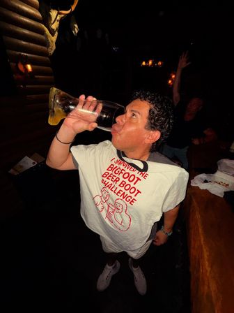 Person wearing a white "I survived the Bigfoot Beer Boot Challenge" shirt chugs from a giant glass beer boot in a dimly lit rustic bar, lively party vibe