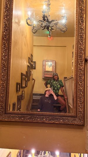 Two friends posing for a mirror selfie on a narrow indoor stairwell, ornate gold-framed mirror reflecting a chandelier, framed photos on textured yellow walls and a wooden banister