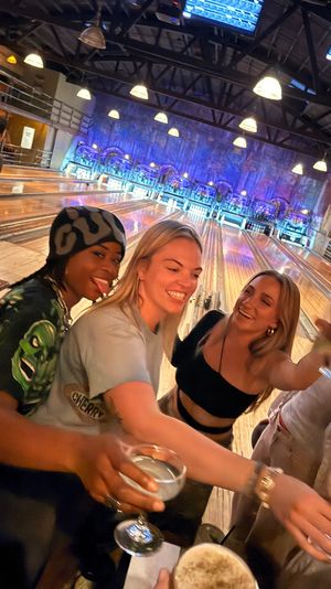 Three friends laughing and toasting with drinks at a lively bowling alley, illuminated lanes and overhead score screens in the background.