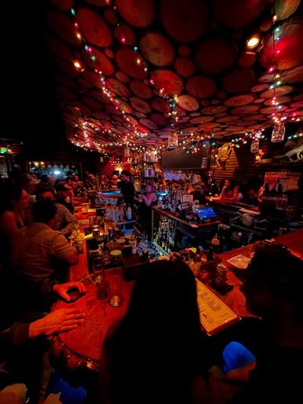 Cozy wood‑paneled bar interior with round log‑slice ceiling, colorful string lights, busy wooden counter, bartenders, bottles and menus—vibrant nightlife scene.