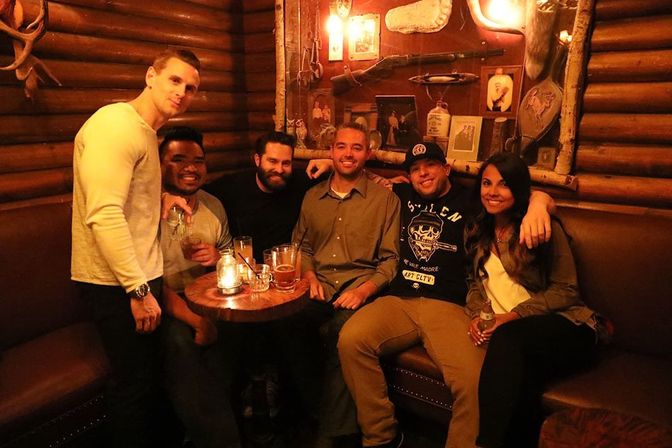 Six friends smiling in a cozy rustic log-cabin bar booth, gathered around a small table with cocktails and candles for a warm night out