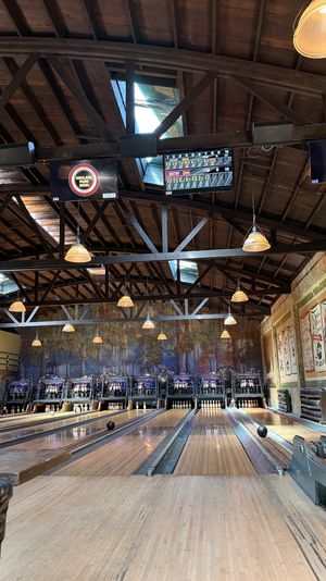 Vintage indoor bowling alley with high exposed-wood beams, hanging pendant lights, electronic score displays, mural backdrop and empty wooden lanes with pins.