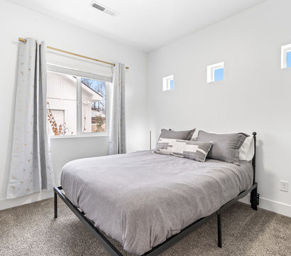 Bright modern bedroom with white walls, gray bedding on a black metal frame, patterned curtains on a gold rod, three small square clerestory windows and plush carpet