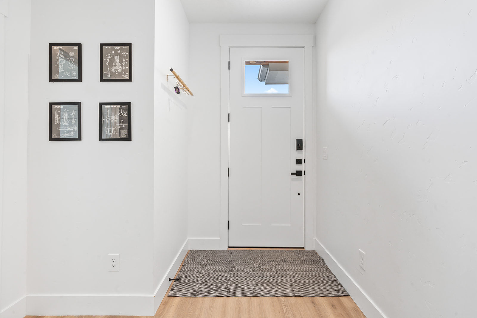 Bright minimalist entryway with a white front door and small window, light wood floor and woven gray rug; left wall shows four black-framed blueprint prints and a wooden coat hook with keys.