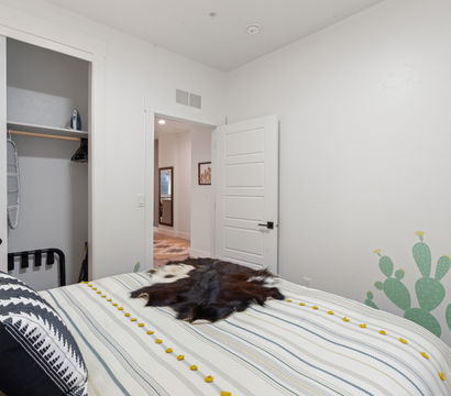 Bright modern bedroom interior with white walls, open closet and hallway doorway; striped bedding with yellow pom‑pom trim, dark cowhide throw and green cactus wall decal.