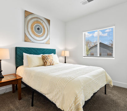 Sunlit modern bedroom with teal velvet headboard, cream textured bedding, patterned accent pillow, circular abstract wall art, matching bedside lamps, window blinds and a leaning full-length mirror.