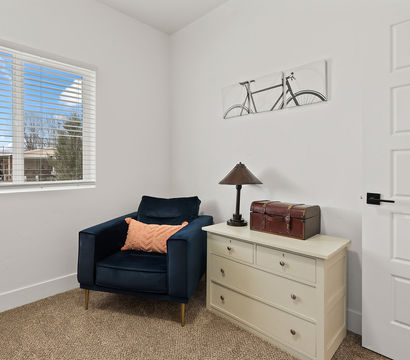 Cozy bright reading nook in a small room with a navy velvet armchair and coral throw pillow, cream three-drawer dresser topped with a vintage leather trunk and table lamp, bicycle wall art, carpeted floor and a window with white blinds.