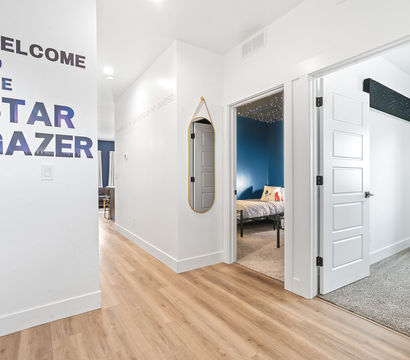 Bright modern hallway with light wood floors and a large "WELCOME TO THE STAR GAZER" wall decal, oval hanging mirror, and open doors to a blue-accent kids' bedroom with a starry ceiling and an adjacent carpeted room.