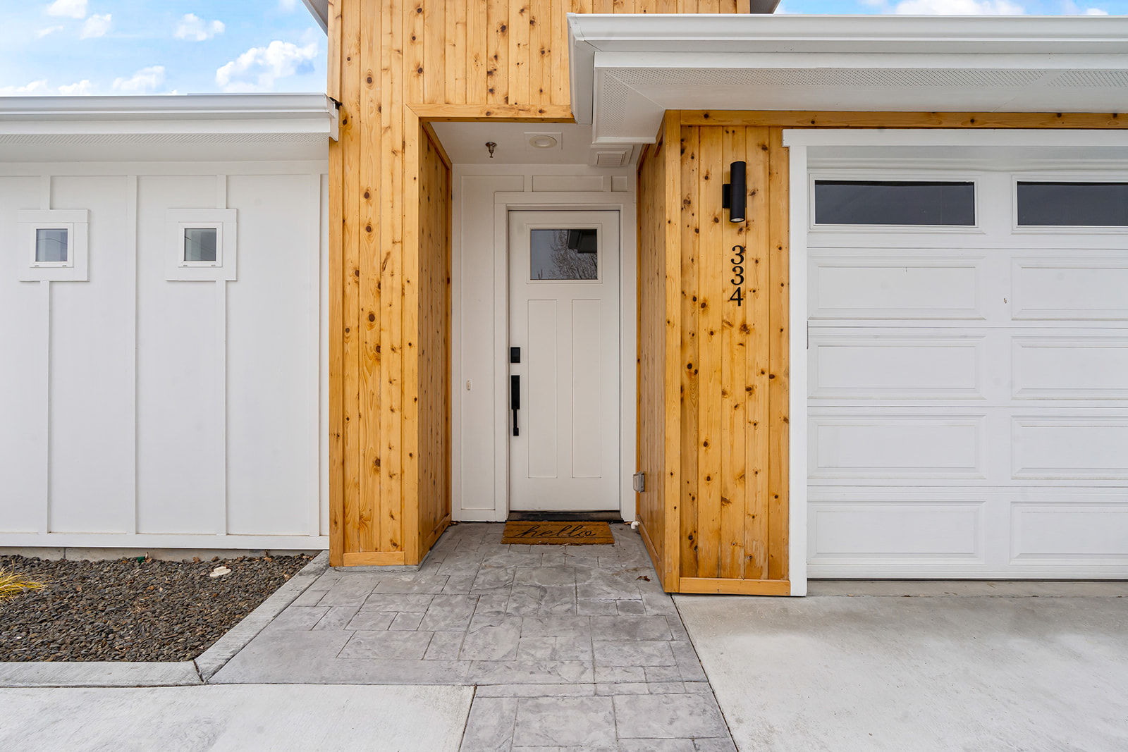 Modern suburban front entry with white paneled door and garage, warm vertical cedar siding, black wall sconce and a welcome "hello" doormat
