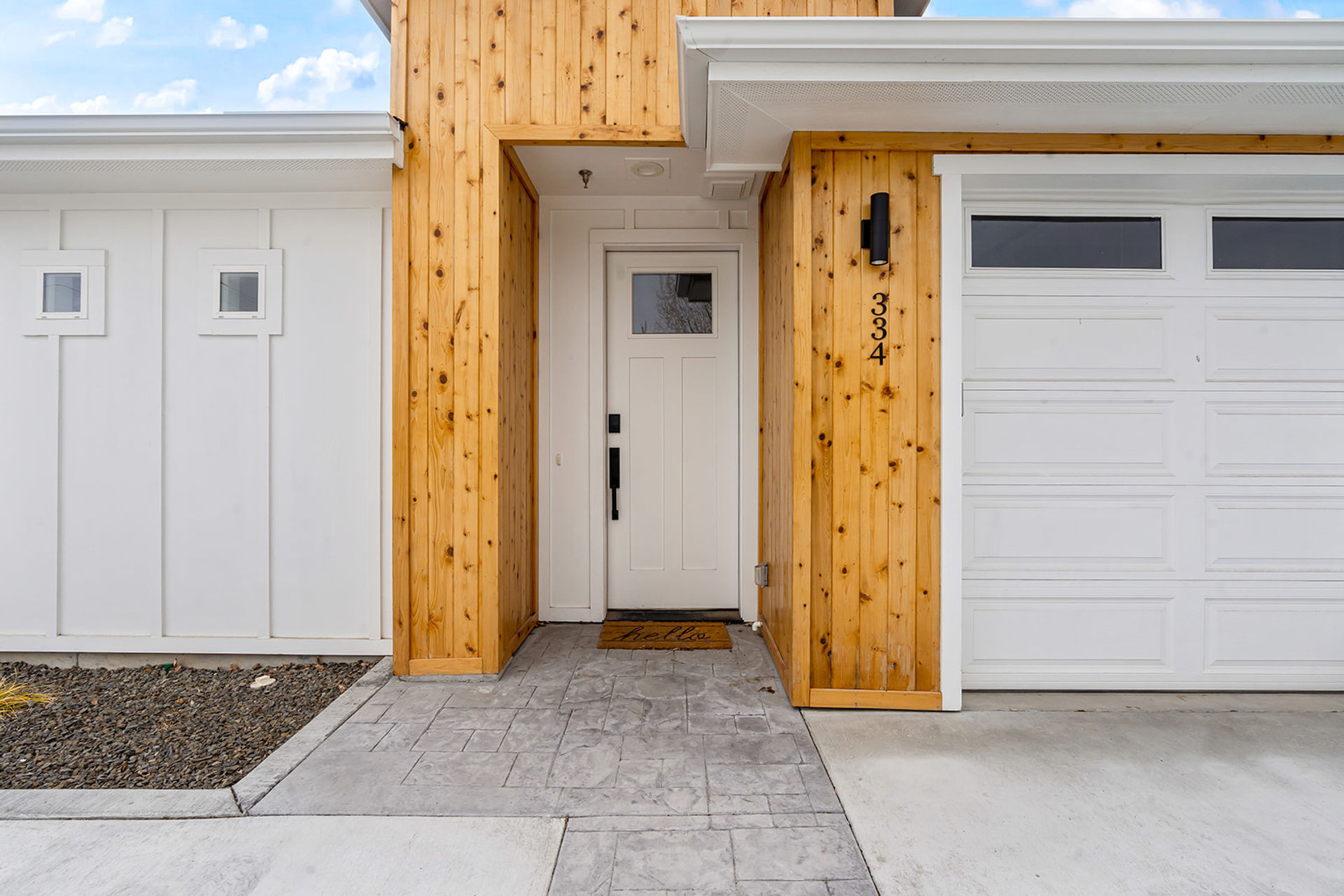 Modern suburban front entry with white paneled door and garage, warm vertical cedar siding, black wall sconce and a welcome "hello" doormat