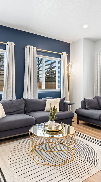 Sunlit modern living room interior with a navy accent wall, two gray sofas, white patterned curtains, gold geometric coffee table and light hardwood floors.