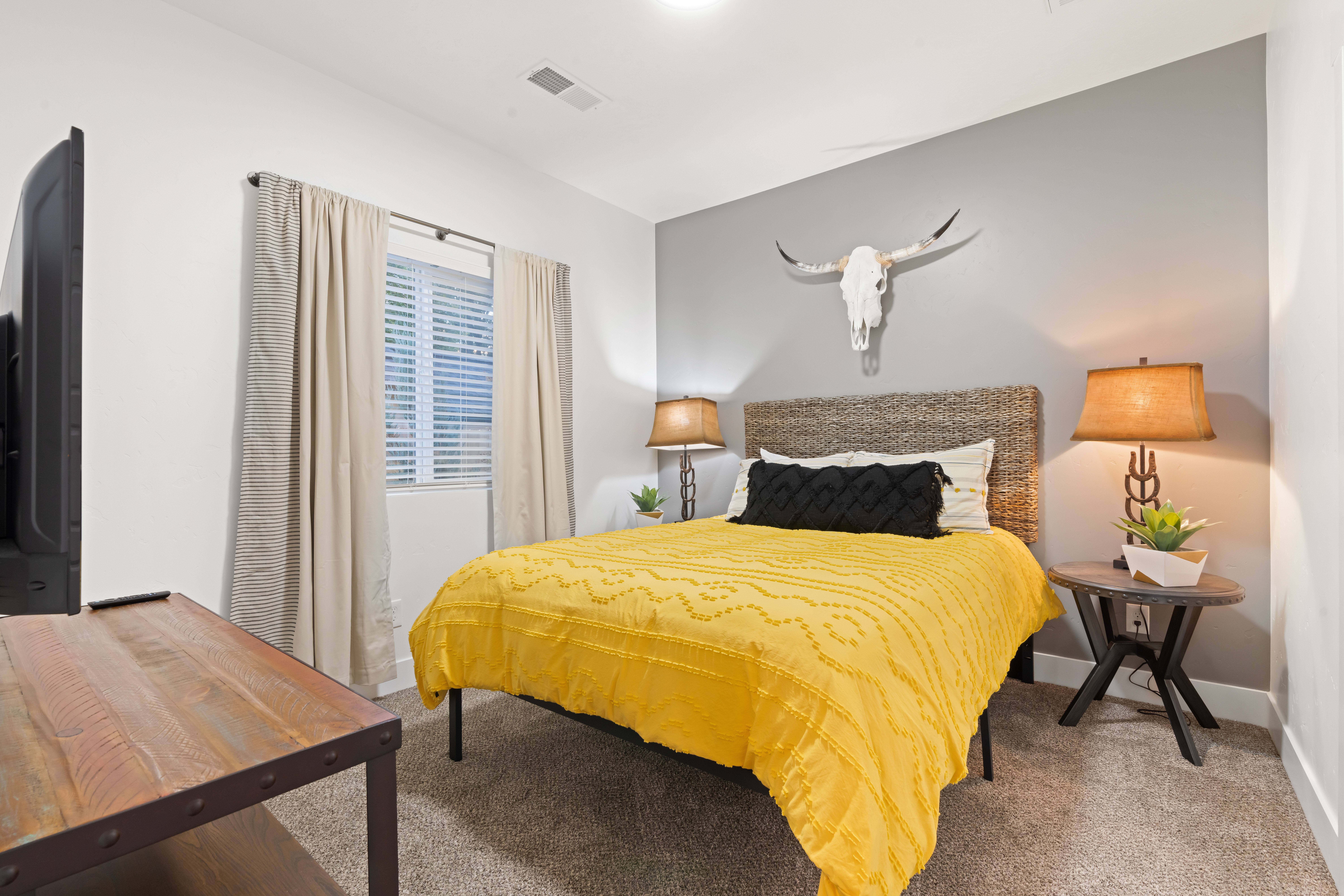Cozy modern bedroom with a sunny yellow bedspread, woven headboard, decorative longhorn skull on a gray accent wall, twin bedside lamps and a curtained window with blinds.