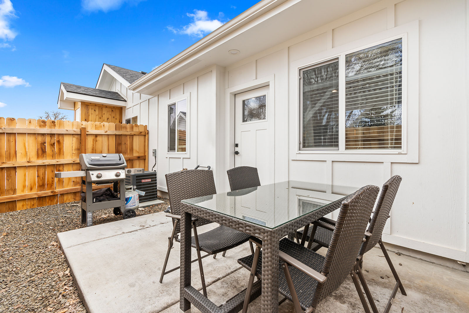 Backyard patio at a modern white-sided home with a glass-top wicker dining table and four chairs, a gas grill beside a wooden privacy fence, concrete slab and bright blue sky.