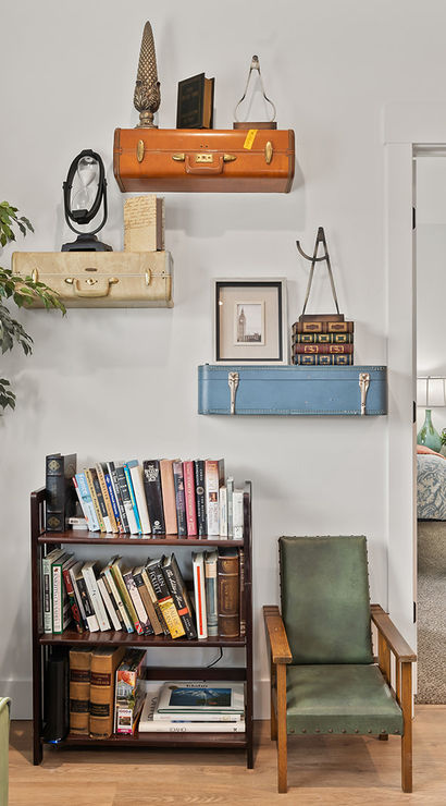 Bright cozy living room reading nook with green armchair, globe, potted ficus, small bookshelf and vintage suitcases mounted as wall shelves, view through doorway into bedroom.
