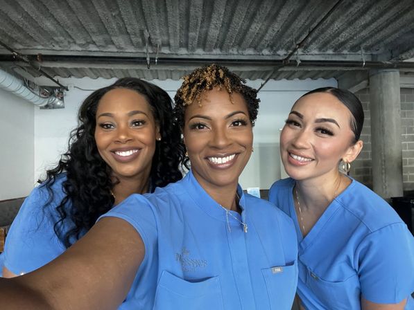 Three smiling healthcare professionals in blue scrubs taking a close-up selfie inside a clinic with exposed concrete ceiling and industrial pipes.