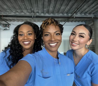 Three smiling healthcare professionals in blue scrubs taking a close-up selfie inside a clinic with exposed concrete ceiling and industrial pipes.