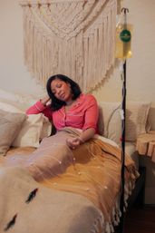 Woman in pink pajamas reclining on a cozy boho bedroom bed under a macramé wall hanging, covered by a tan blanket and connected to an IV drip in a yellow bedside bag.