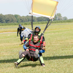 Tandem skydiving landing on a grassy airfield — instructor in a helmet guiding a passenger in a red harness under a yellow parachute canopy.