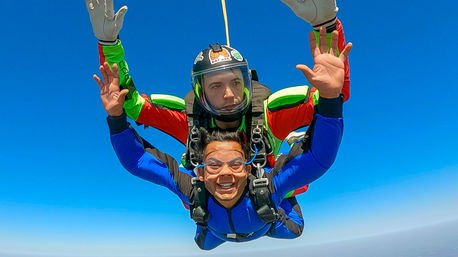 Tandem skydive mid-freefall against a clear blue sky, smiling passenger in a blue jumpsuit and goggles strapped to an instructor in red-and-green gear