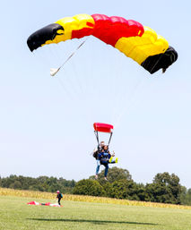 Tandem skydivers landing under a red, yellow and black parachute over a rural grassy field with trees and a cornfield in the background.