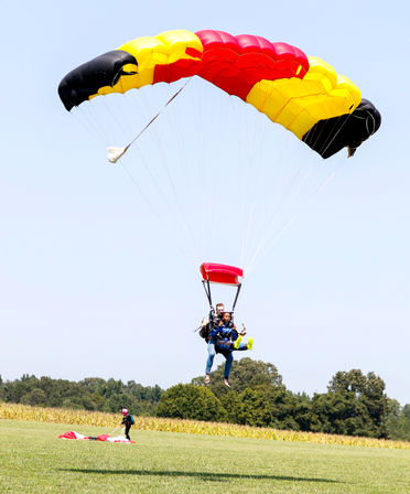 Tandem skydivers landing under a red, yellow and black parachute over a rural grassy field with trees and a cornfield in the background.