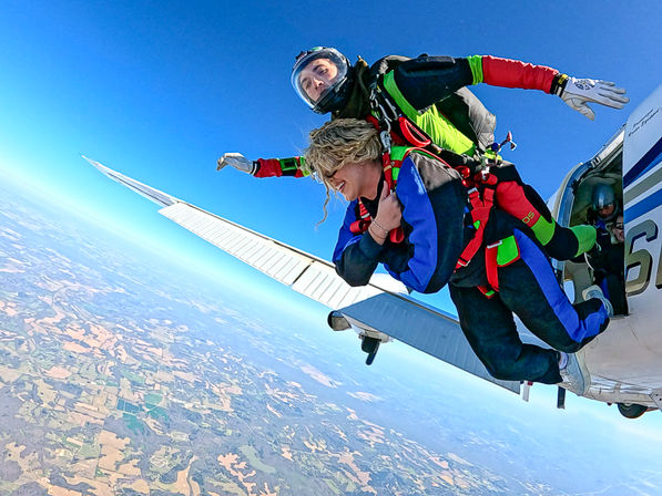 Tandem skydiving pair exiting a small airplane, instructor strapped behind the passenger as they freefall over patchwork farmland beneath a clear blue sky
