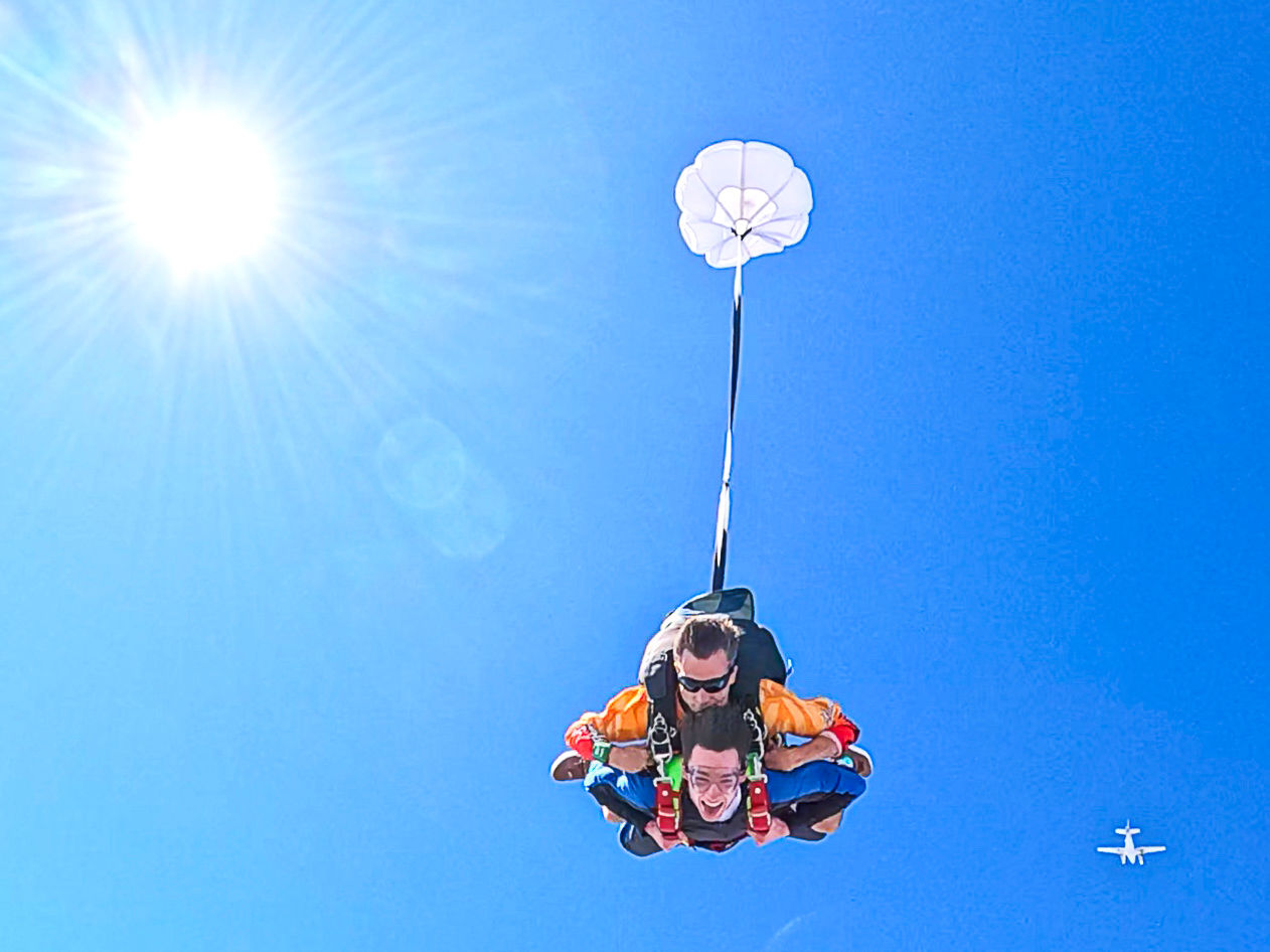 Tandem skydiving pair descending in a clear blue sky with sun flare, small drogue parachute deployed and a distant airplane visible