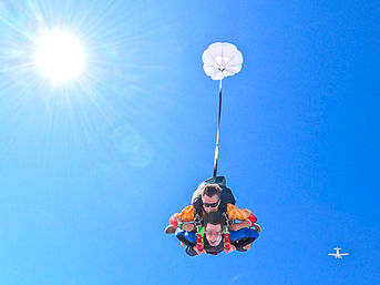 Tandem skydiving pair descending in a clear blue sky with sun flare, small drogue parachute deployed and a distant airplane visible