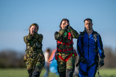 Three skydivers walking across a grassy drop zone after landing — two in camouflage jumpsuits with red harnesses and one in a blue suit under a clear blue sky.
