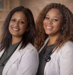 Two smiling women doctors in white coats with stethoscopes, friendly portrait in a warm medical clinic setting