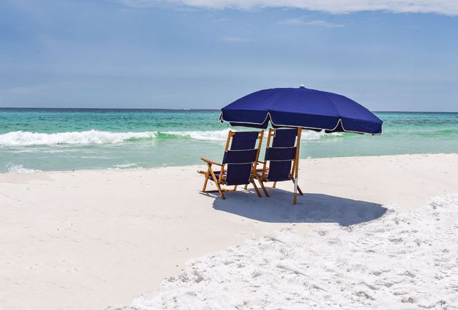 Two wooden lounge chairs under a navy beach umbrella on a white-sand beach with turquoise waves and a calm blue sky — relaxing coastal scene.