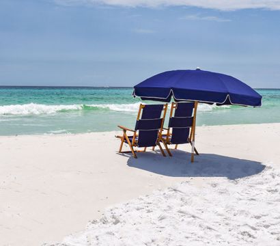 Two wooden lounge chairs under a navy beach umbrella on a white-sand beach with turquoise waves and a calm blue sky — relaxing coastal scene.