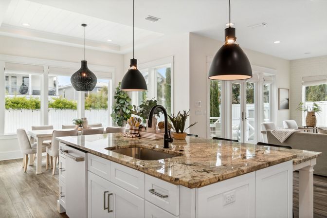 Bright open-concept kitchen with polished granite island, matte-black pendant lights, white cabinetry, dining nook and large windows flooding the space with natural light.