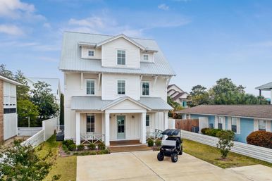 Three-story white coastal beach house with metal roof and front porch, small golf cart parked on concrete driveway, manicured lawn and palm trees in a suburban seaside neighborhood under a blue sky