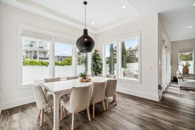 Sunlit modern dining room with a white farmhouse table and six beige upholstered chairs, a black woven pendant light, wood-look flooring, and large windows overlooking a fenced yard and outdoor patio.