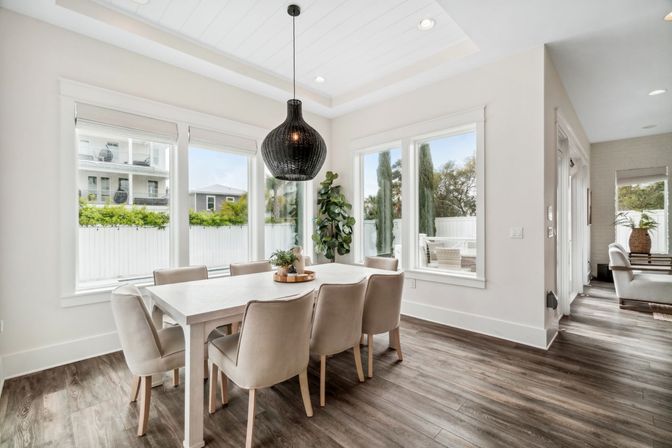 Sunlit modern dining room with a white farmhouse table and six beige upholstered chairs, a black woven pendant light, wood-look flooring, and large windows overlooking a fenced yard and outdoor patio.