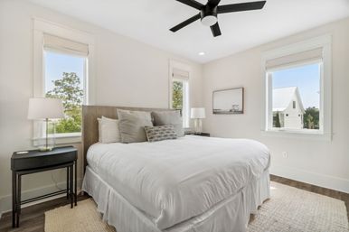 Sunlit modern bedroom with white bedding, wood headboard, neutral throw pillows, glass bedside lamps, black ceiling fan, hardwood floors and three tall windows showing trees and a neighboring white house.