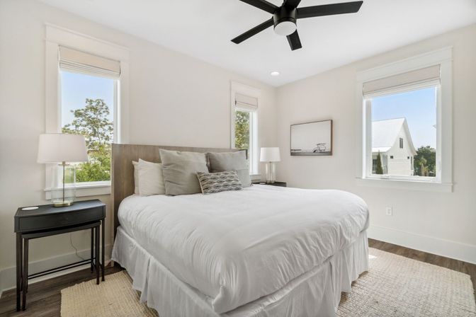 Sunlit modern bedroom with white bedding, wood headboard, neutral throw pillows, glass bedside lamps, black ceiling fan, hardwood floors and three tall windows showing trees and a neighboring white house.