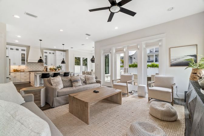 Bright coastal-style open-concept living room and kitchen with white cabinets, black pendant island lights, beige sofa, wood coffee table, woven rug and glass doors to a sunny patio