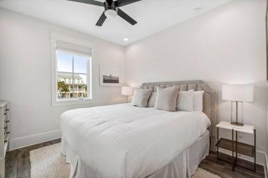 Bright modern bedroom with white bedding and gray upholstered headboard, bedside lamps, ceiling fan, hardwood floors and a window view of neighboring homes.
