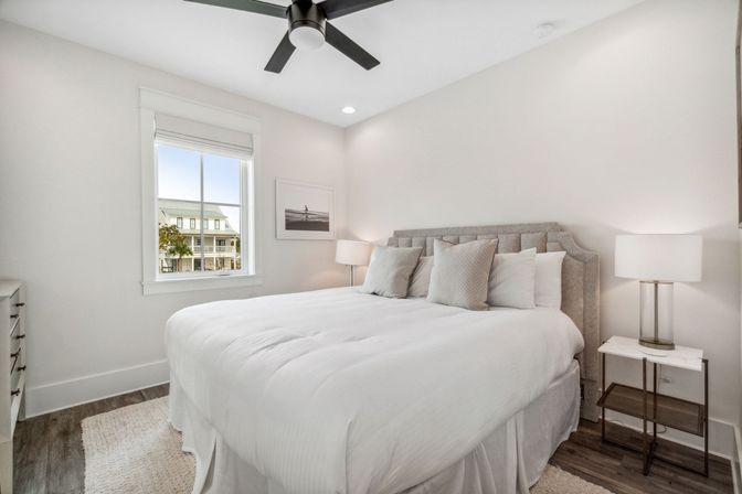 Bright modern bedroom with white bedding and gray upholstered headboard, bedside lamps, ceiling fan, hardwood floors and a window view of neighboring homes.