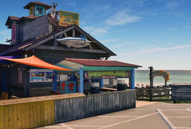 Colorful beachfront seafood shack on a wooden boardwalk with a large mounted marlin, orange canopy, outdoor counter and calm ocean beach in the background