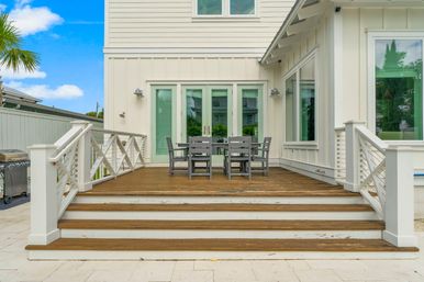 Coastal-style white house deck with wooden steps and cable railing, gray outdoor dining table and chairs in front of mint-green French doors, grill to the side and palm tree under a blue sky