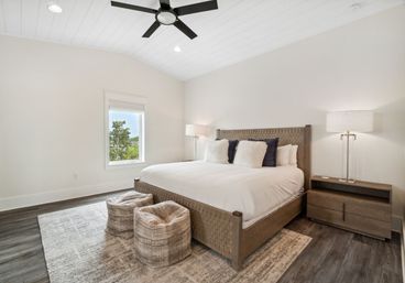 Cozy bright modern bedroom with vaulted white shiplap ceiling and black ceiling fan; woven wood king bed with white bedding and fluffy pillows, two textured poufs on a neutral rug, wooden nightstand and lamp, window with tree view.